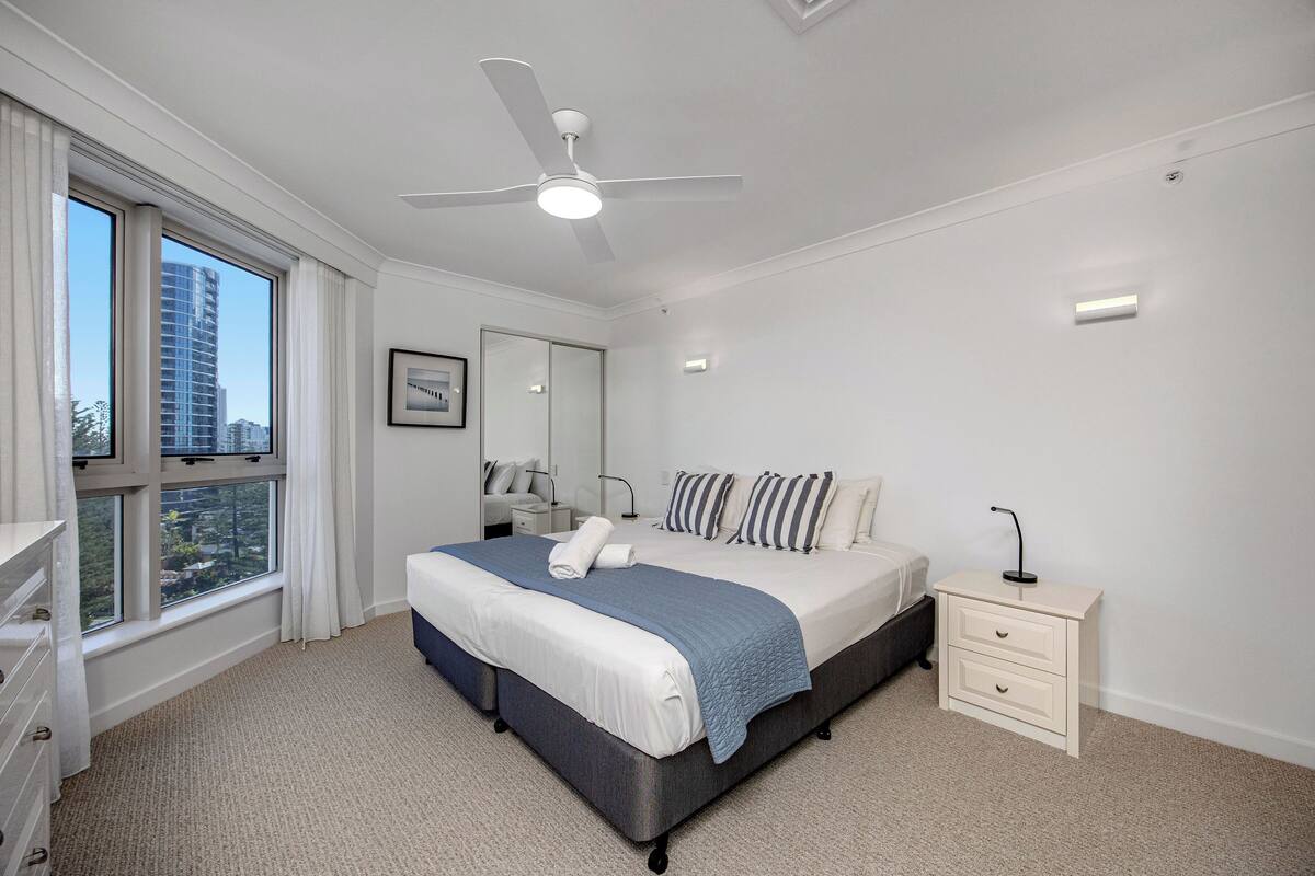 A bedroom with a large bed, a nightstand, a ceiling fan, and a window with curtains.