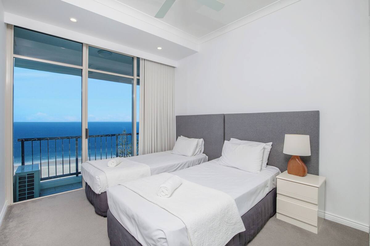 A hotel room with two beds, a nightstand with a lamp, and a balcony with a view of the ocean.