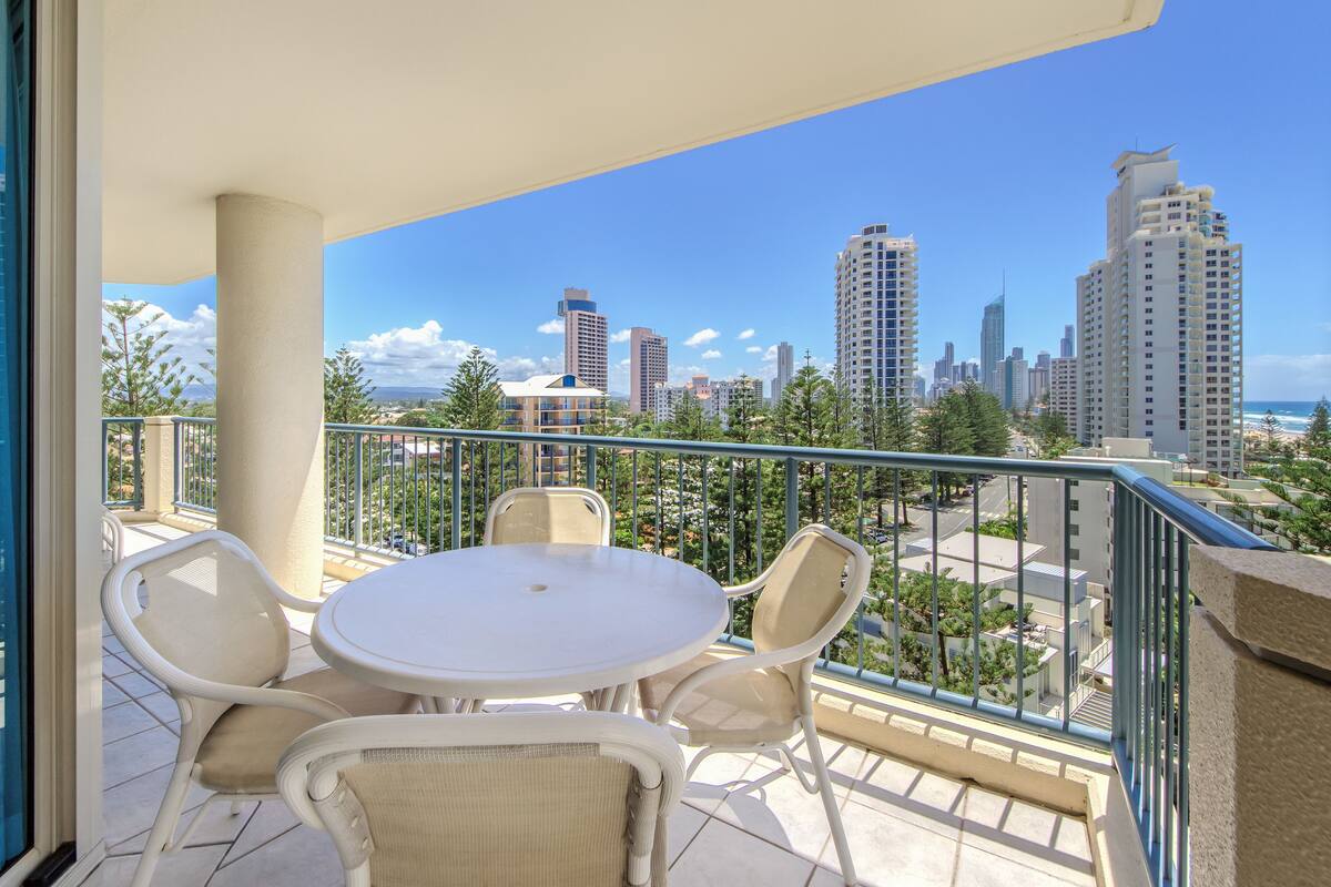 A balcony with a white table and chairs, offering a view of a city skyline and beach.