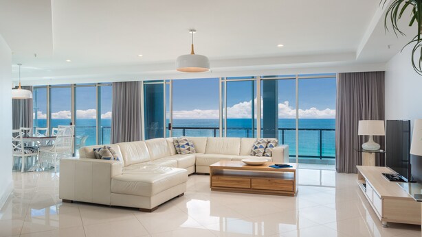 A modern living room with a large sectional sofa, a coffee table, and a TV stand. The room has large windows offering a view of the ocean.