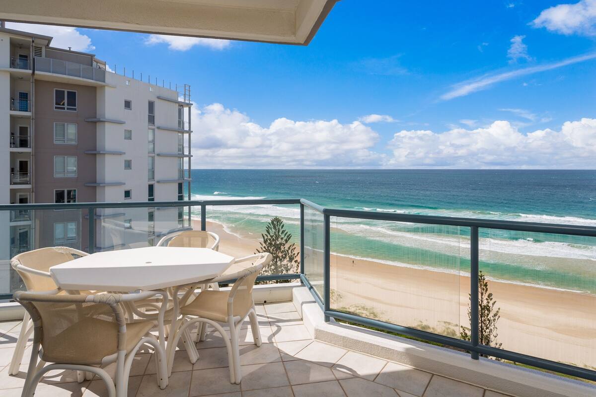 A balcony with a table and chairs overlooking a beach and ocean.