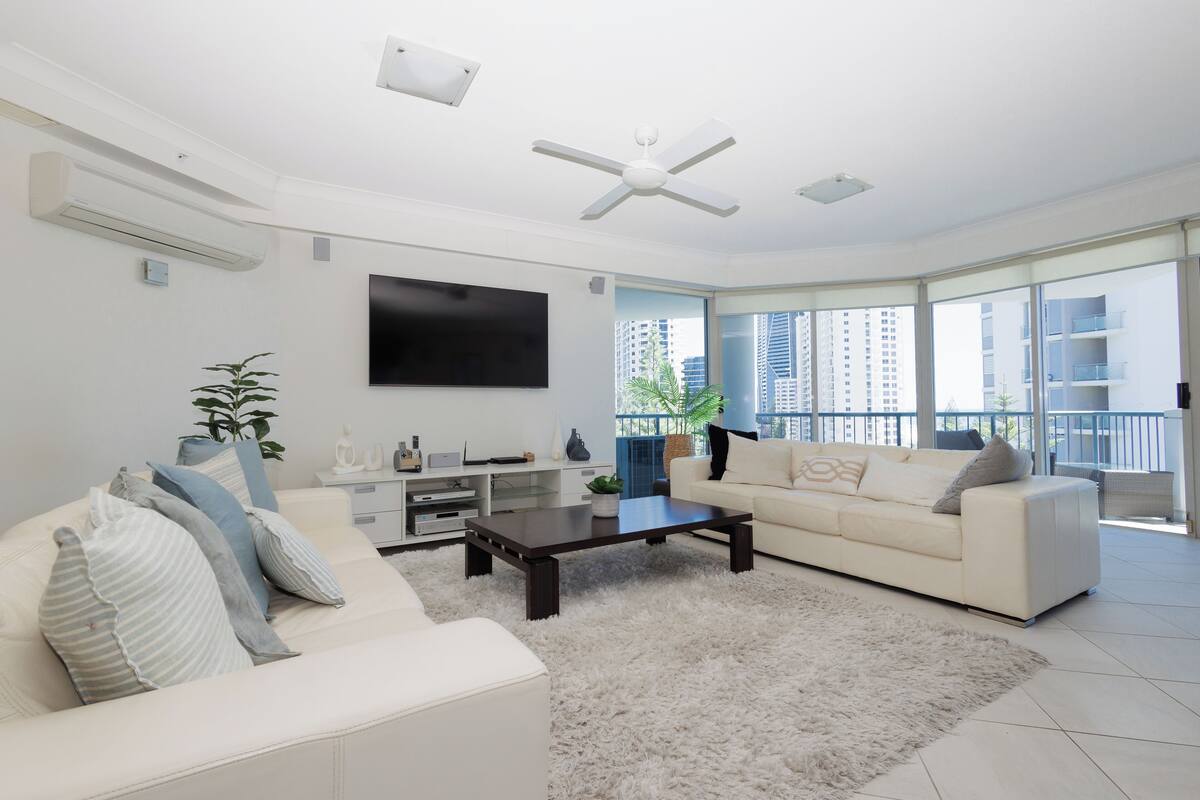 A modern living room with a white sofa, a coffee table, and a flat-screen TV. There is a large window with a view of tall buildings.