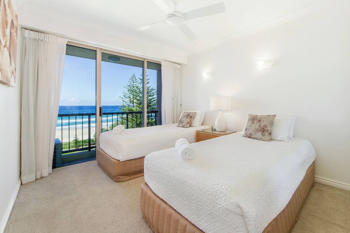 A hotel room with two beds, a ceiling fan, a view of the ocean, and a balcony with a railing.
