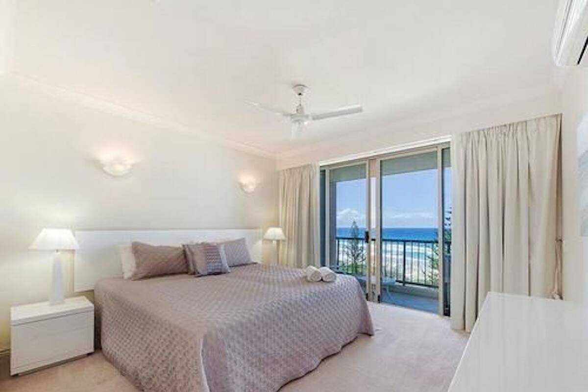 A hotel room with a large bed, a ceiling fan, a nightstand, and a view of the ocean through a balcony.