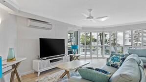 A modern living room with a flat-screen TV, a ceiling fan, and large windows with blinds.