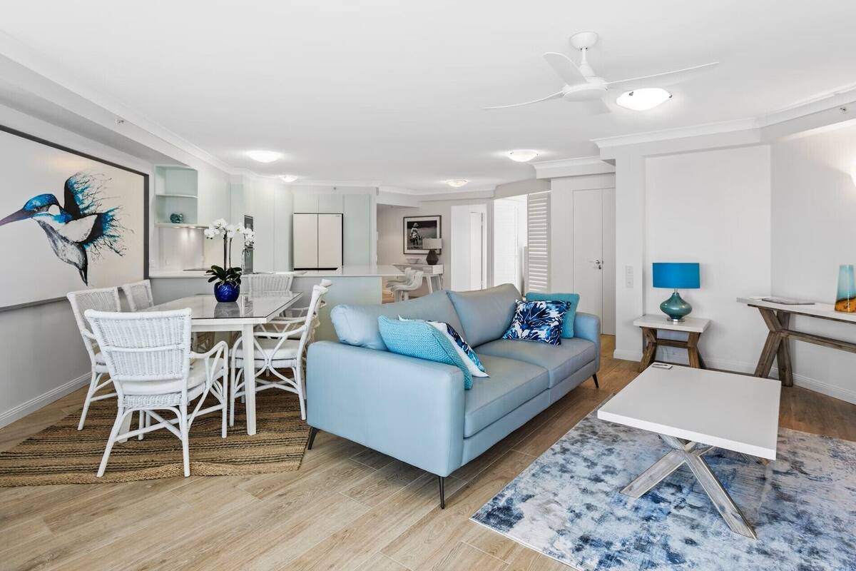 A modern living room with a light blue sofa, white wicker chairs, a dining table, and a ceiling fan.
