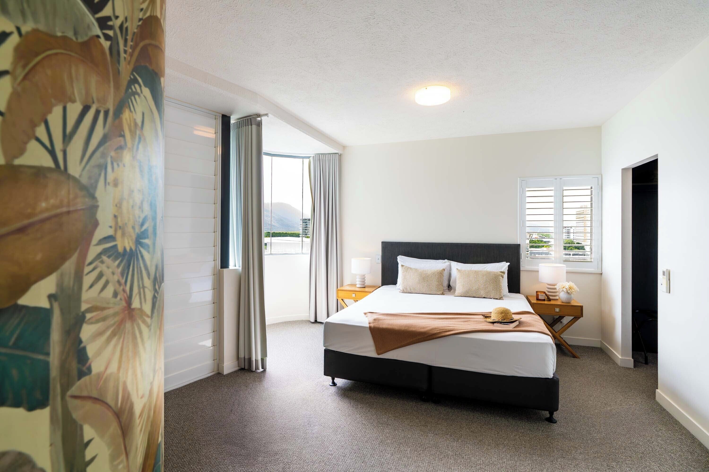 A bedroom with a large bed, a nightstand, a window with blinds, and a view of greenery outside.