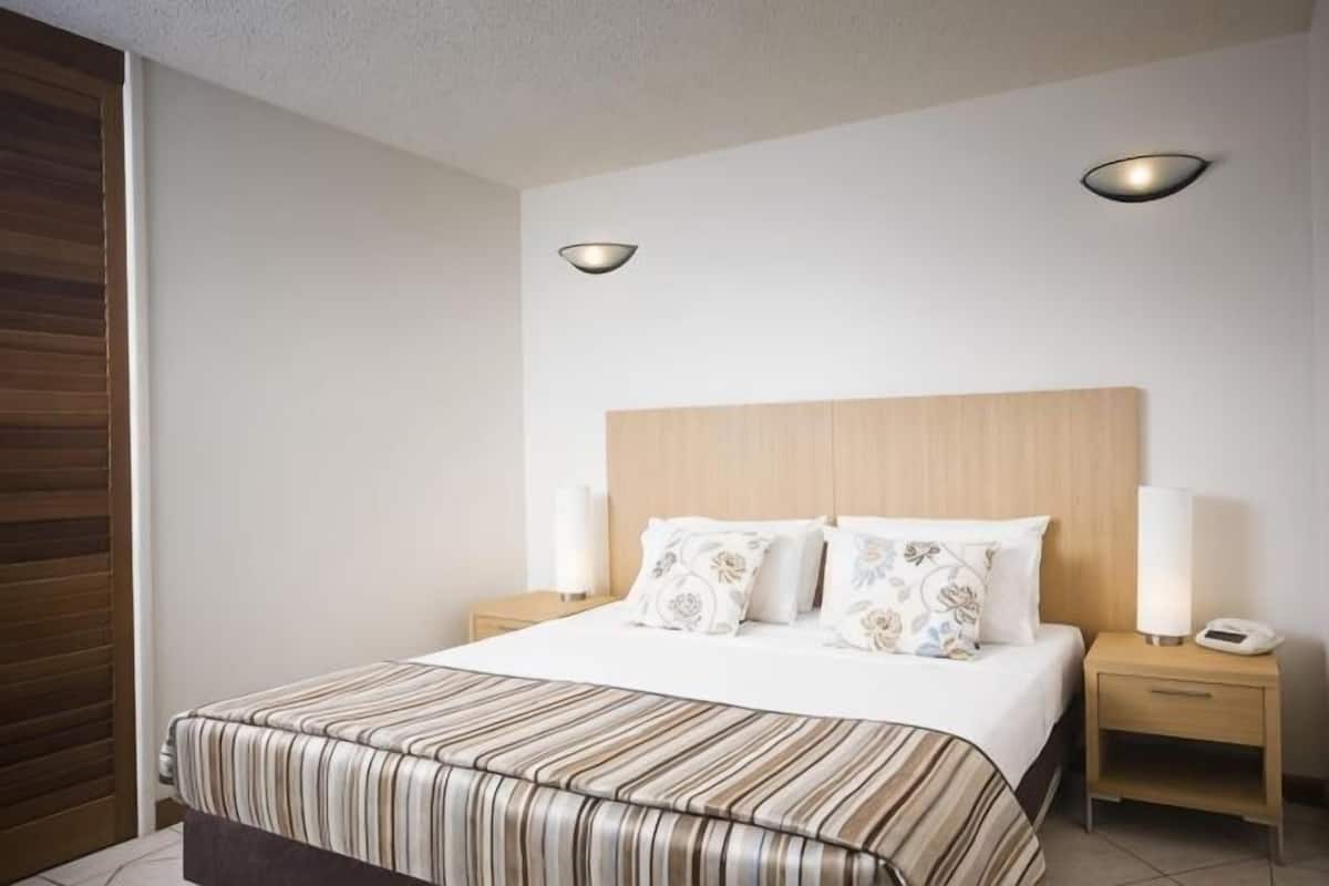 Una habitación de hotel con una cama grande, dos mesitas de noche, un cabecero de madera y dos lámparas fijadas a la pared.