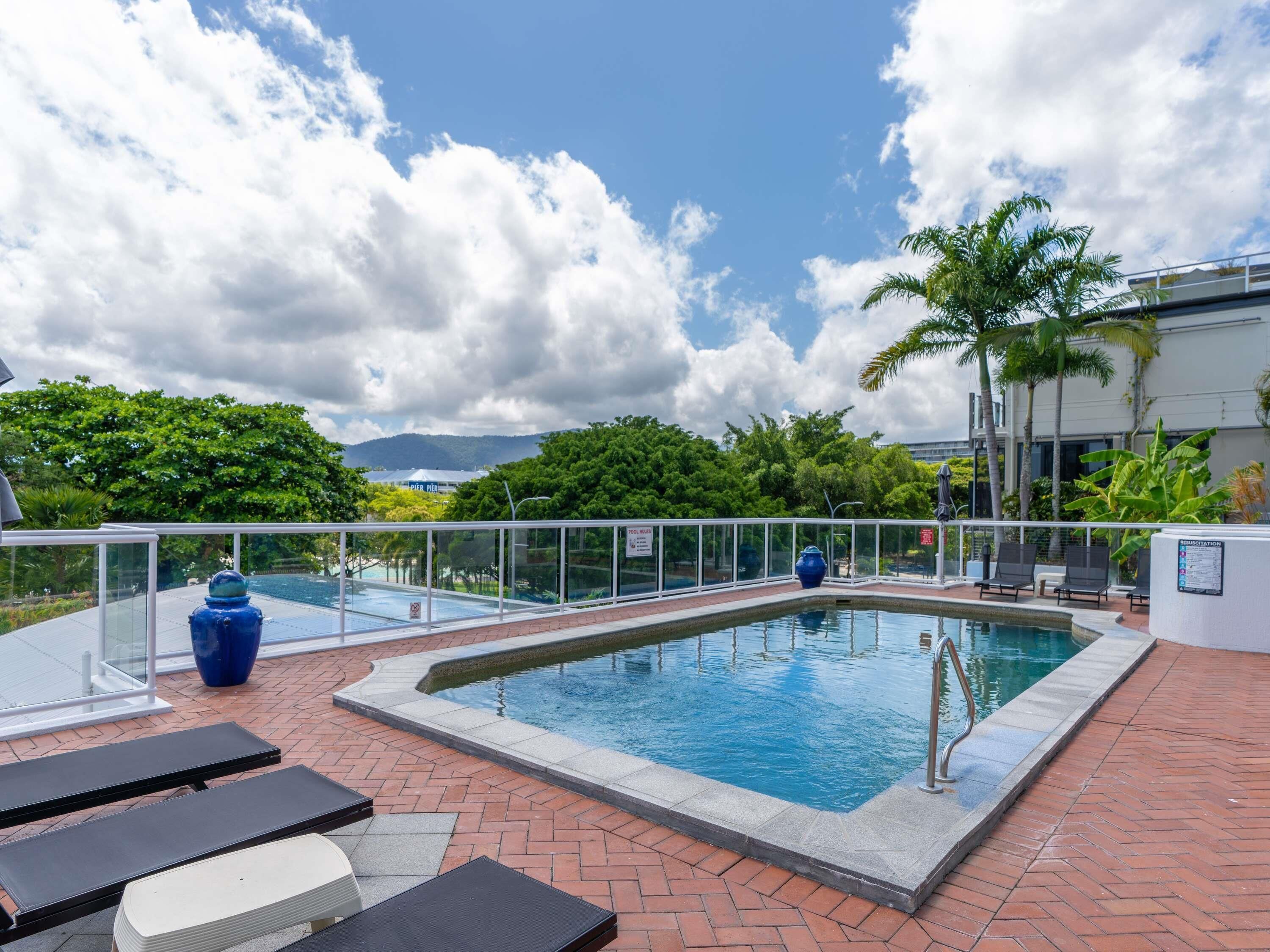 A rooftop pool with lounge chairs, a blue vase, and a view of a building and trees.