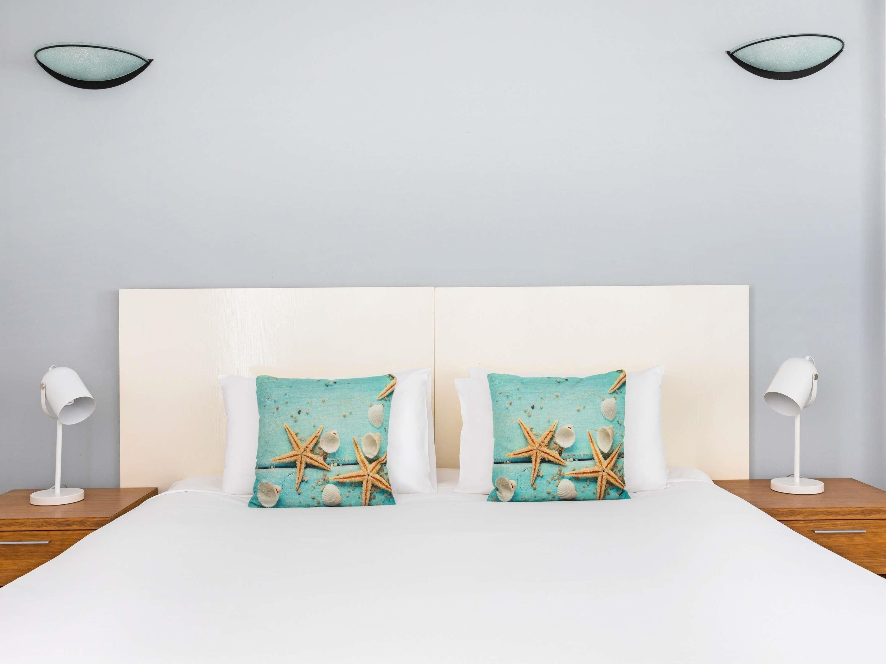 A double bed with white bedding, two matching pillows with starfish and seashells, a white headboard, and two bedside lamps on wooden nightstands.