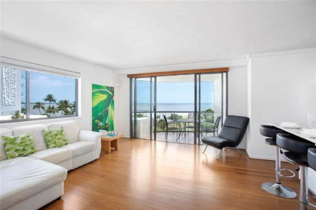 A spacious living area with a white sofa, a green abstract painting, a dining table with chairs, and a balcony with a view of the ocean.