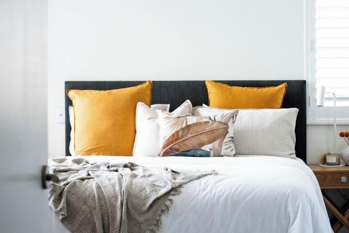 A bed with a dark headboard, white bedding, and two mustard-colored pillows.