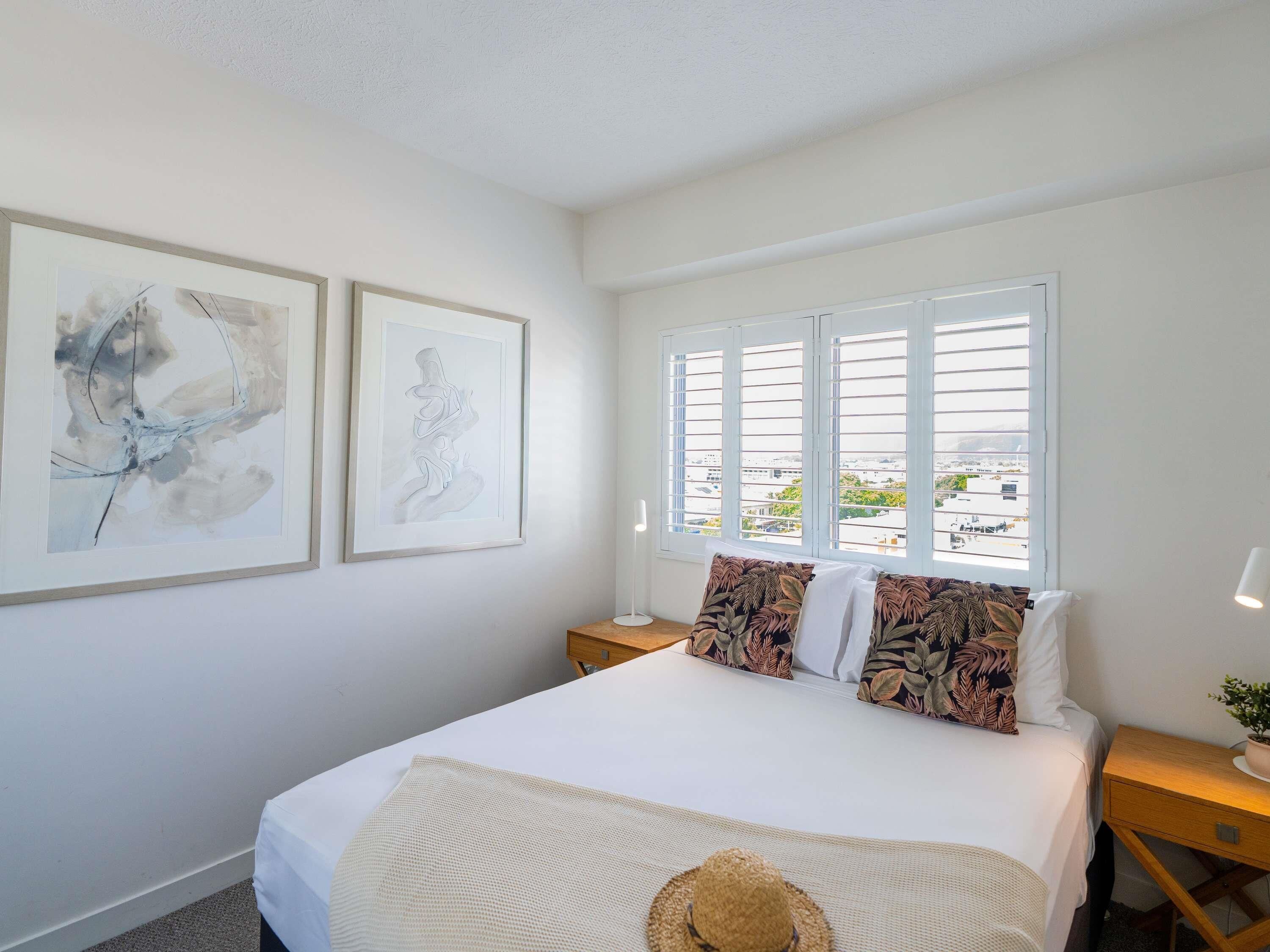 A bedroom with a bed, two framed abstract artworks, a window with blinds, and a nightstand with a lamp.