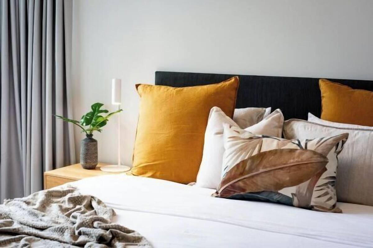 A neatly made bed with multiple pillows, a bedside plant, and a bedside lamp.