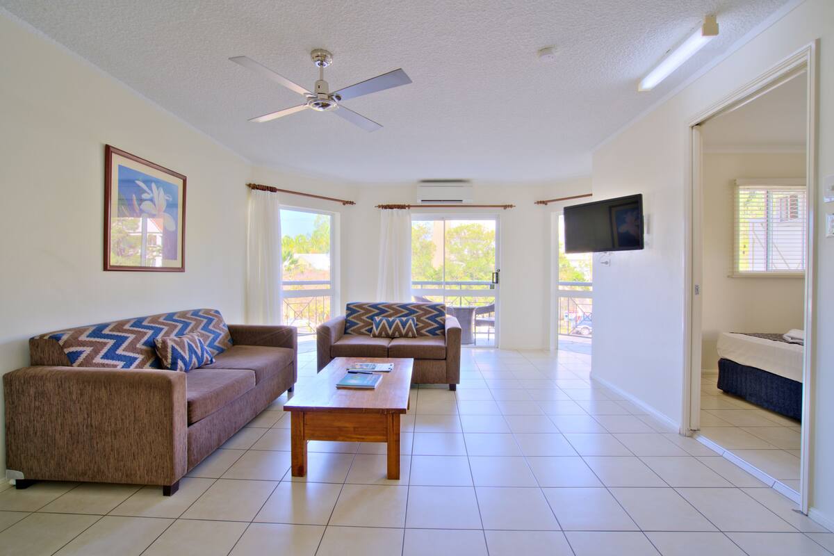 A spacious living room with two sofas, a coffee table, a ceiling fan, and a TV mounted on the wall.
