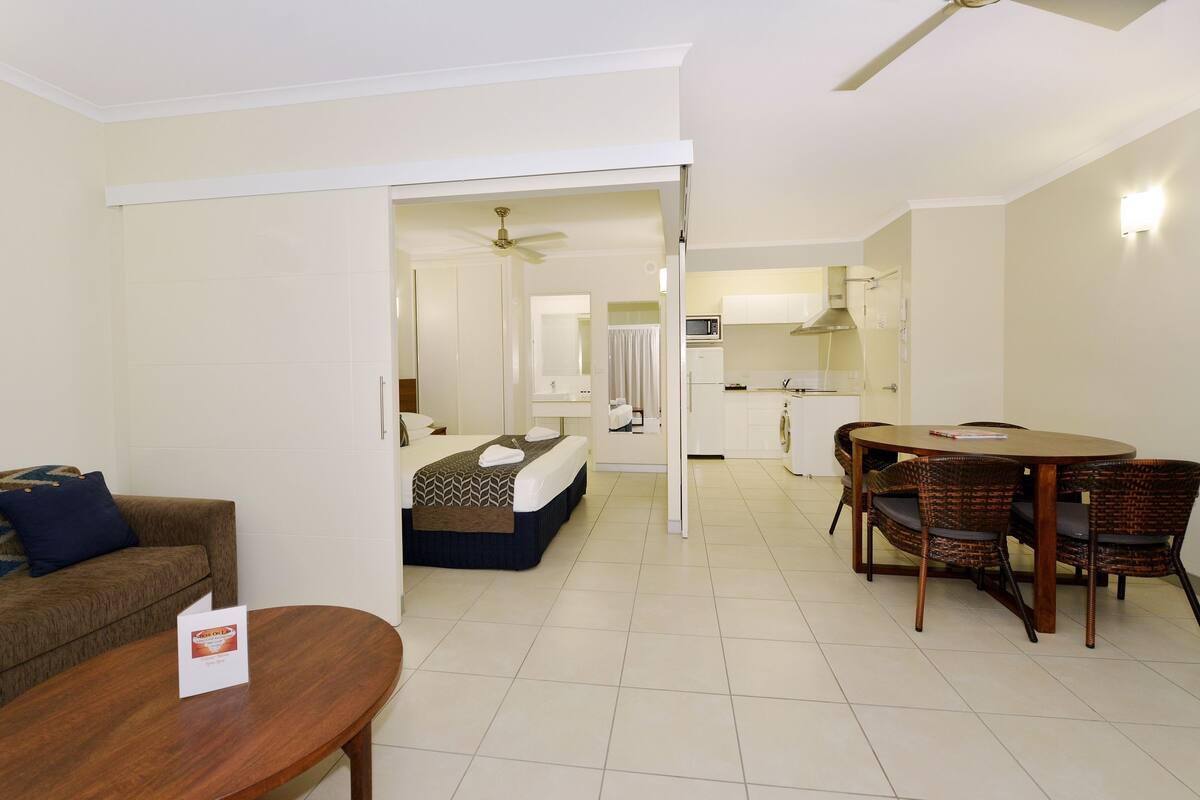 A hotel room with a bed, a dining table with chairs, a kitchenette, and a bathroom visible through an open door.