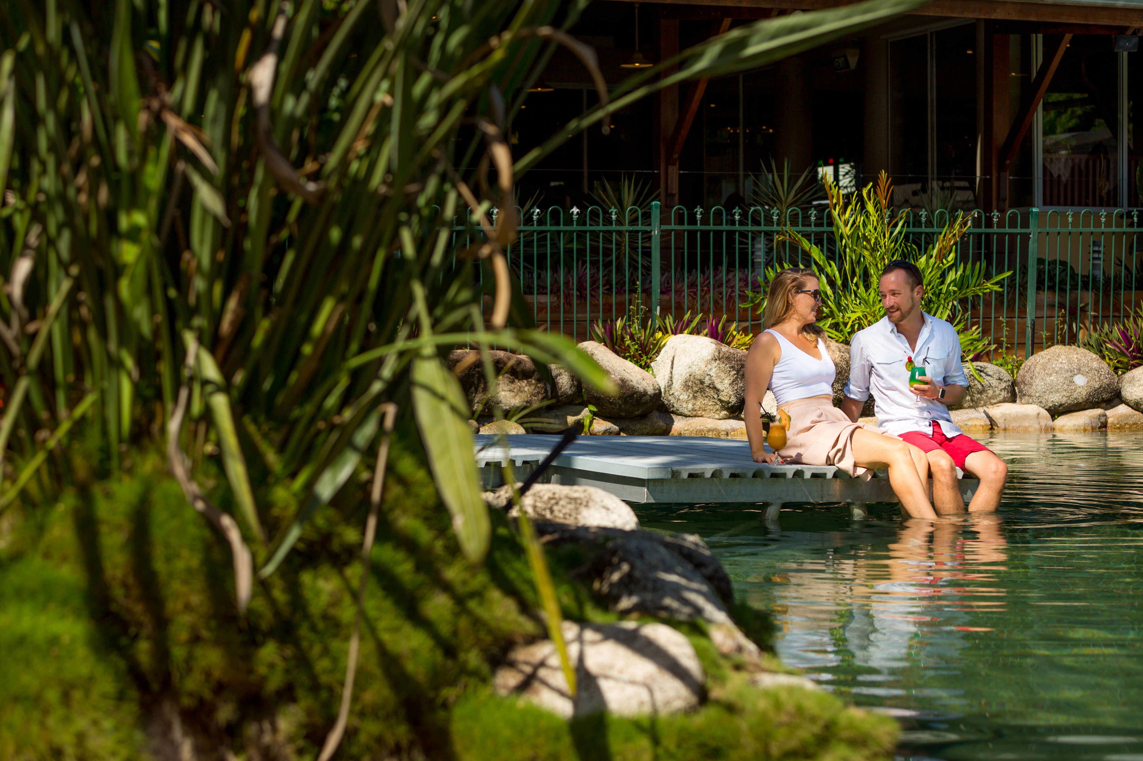 A couple sitting on a floating platform in a pool surrounded by rocks and greenery.