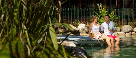 A couple sitting on a floating platform in a pool surrounded by rocks and greenery.