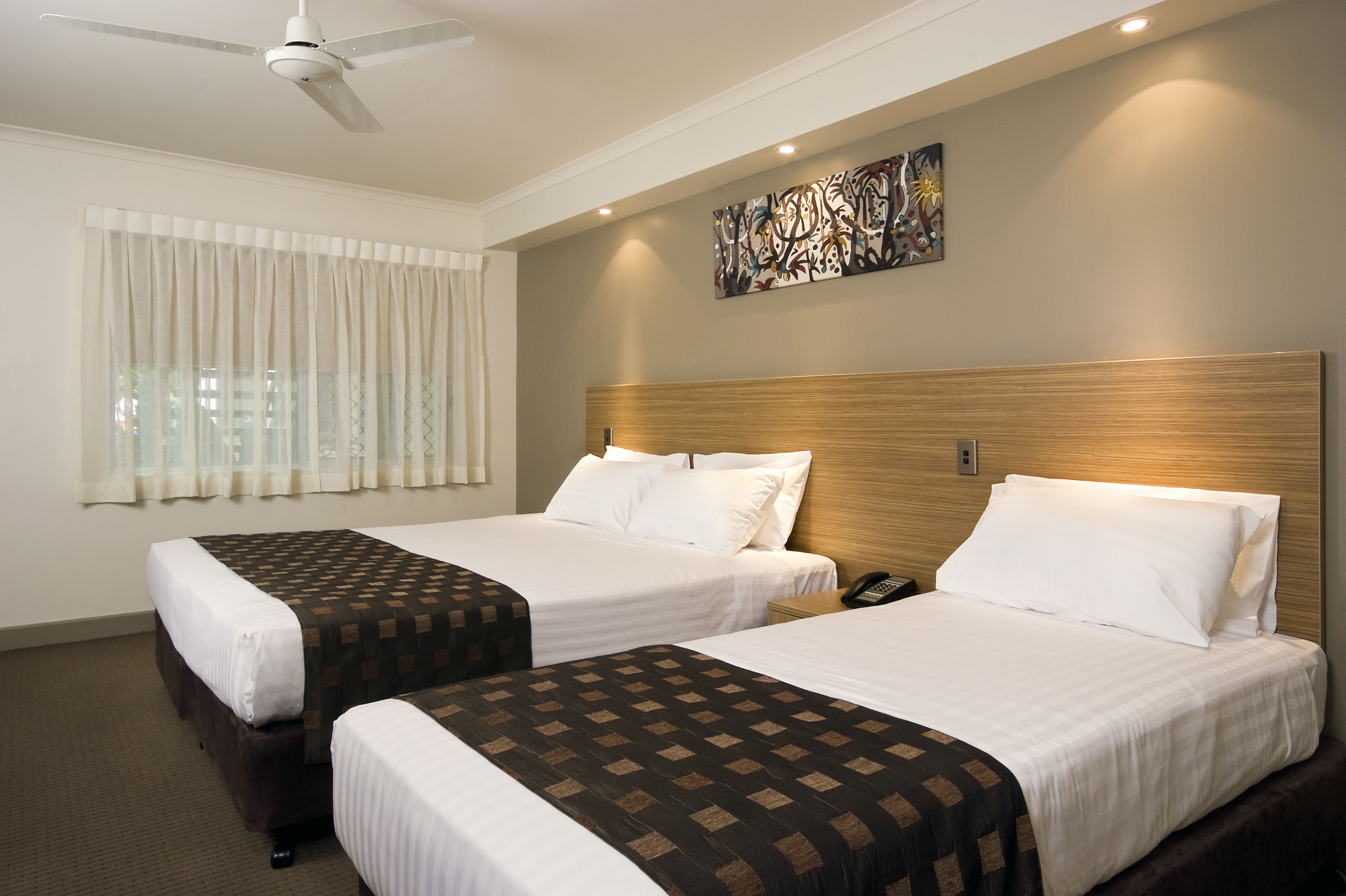 Habitación de hotel con dos camas, un ventilador de techo, un ventanal con cortinas y una obra de arte colgada en la pared.