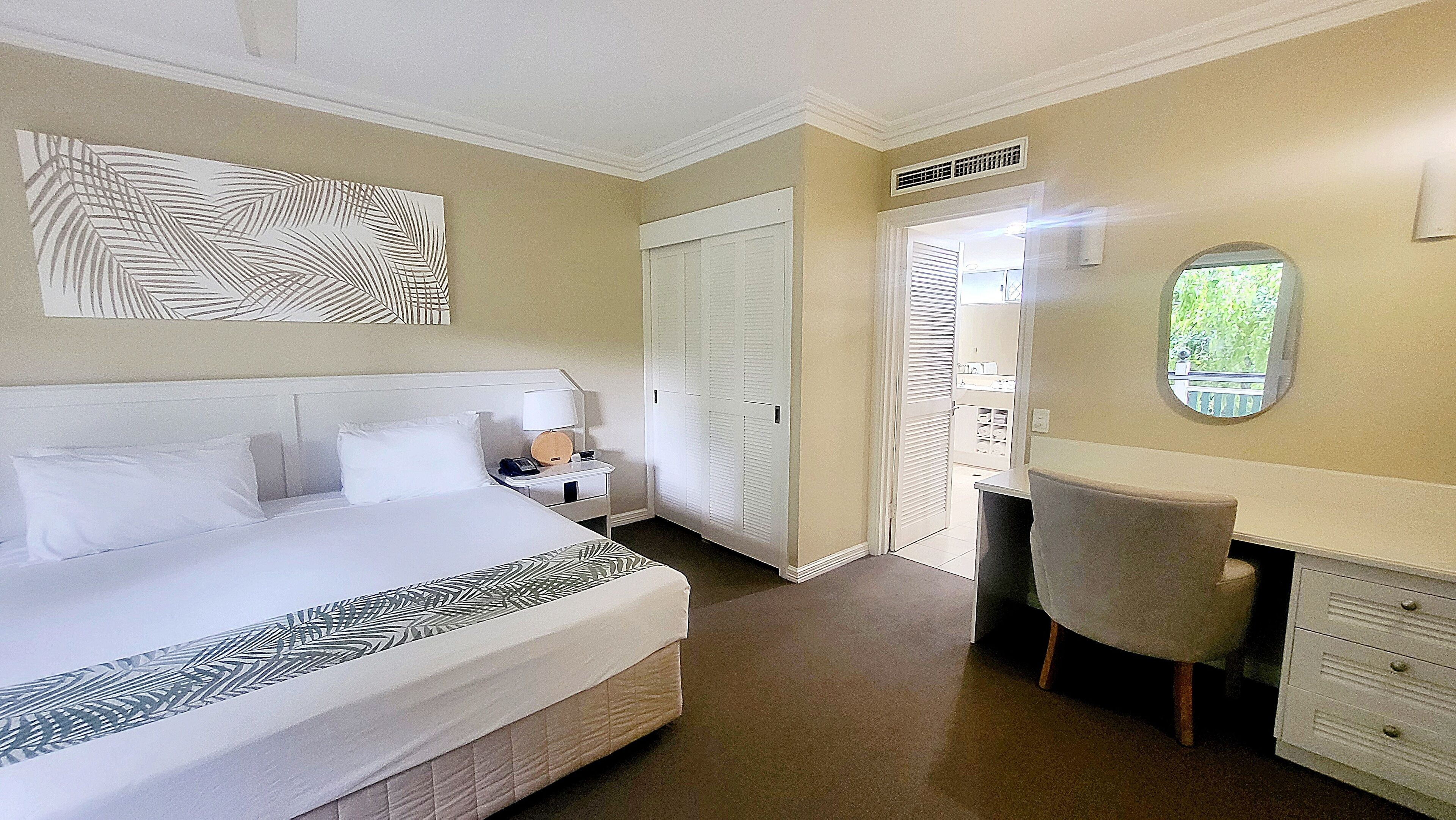 A hotel room with a large bed, a desk, and a chair. There is a wardrobe and a mirror on the wall.