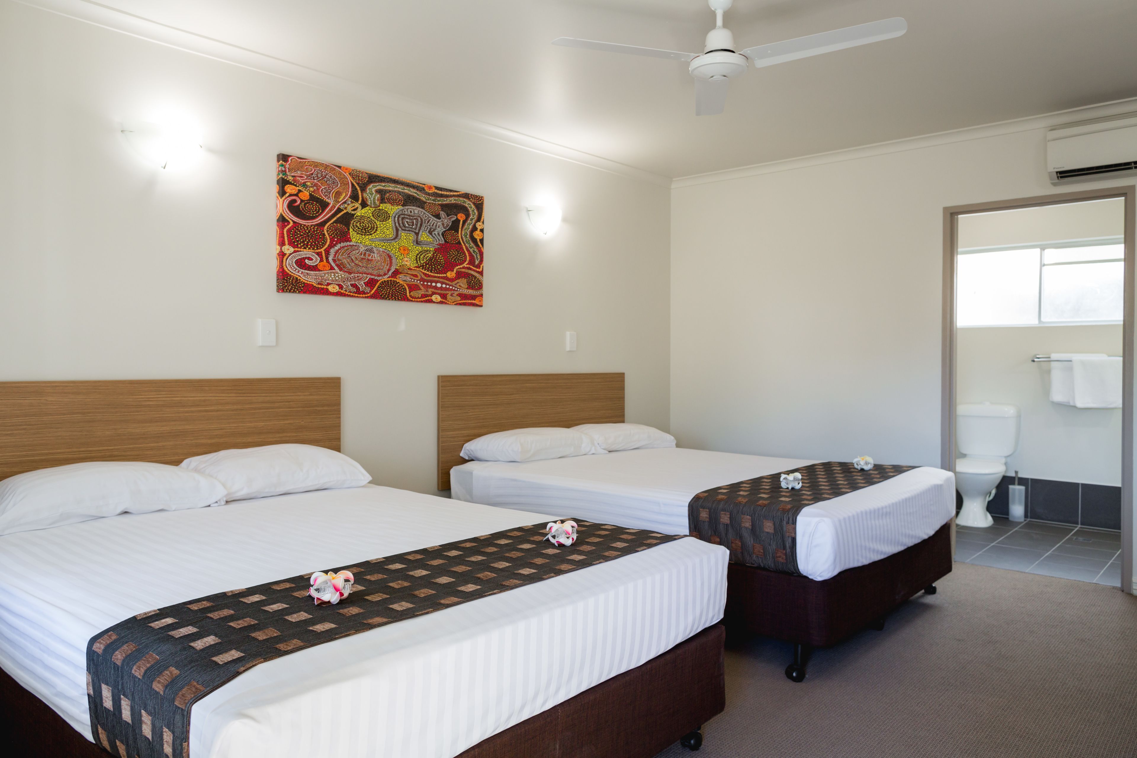 A hotel room with two single beds, a ceiling fan, a colorful artwork on the wall, and a bathroom with a toilet and sink.