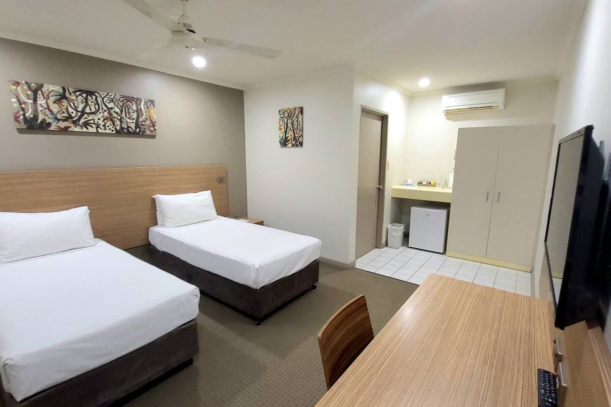 A hotel room with two single beds, a wooden desk with a chair, a television, and artwork on the walls.
