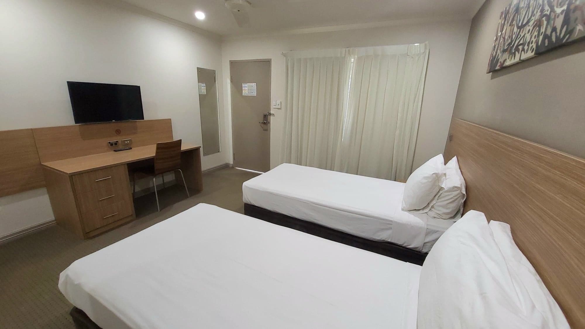 A hotel room with two single beds, a wooden desk with a chair, a television, and a door with a sign.
