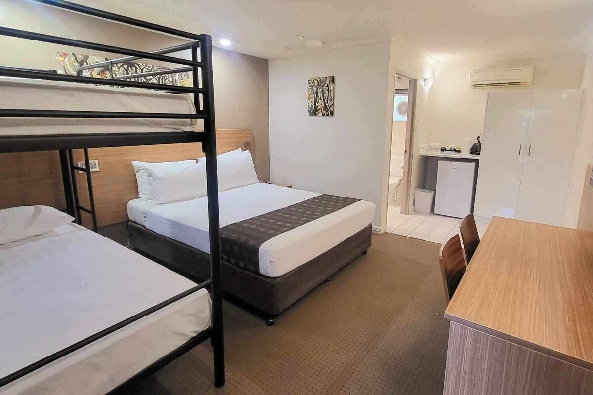 A hotel room with bunk beds, a single bed, a desk, a chair, a television, and a small kitchenette.