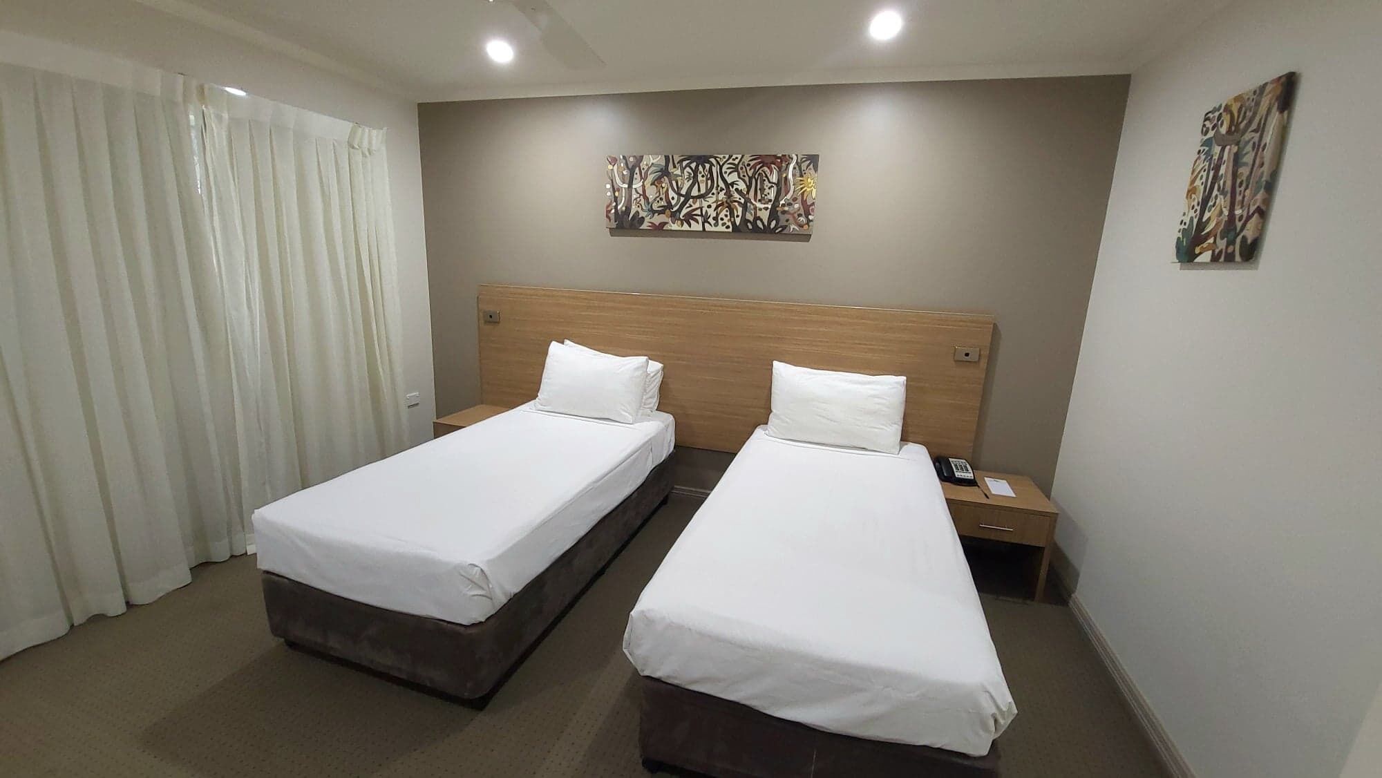 A hotel room with two single beds, a wooden headboard, a nightstand with a phone, and abstract wall art.