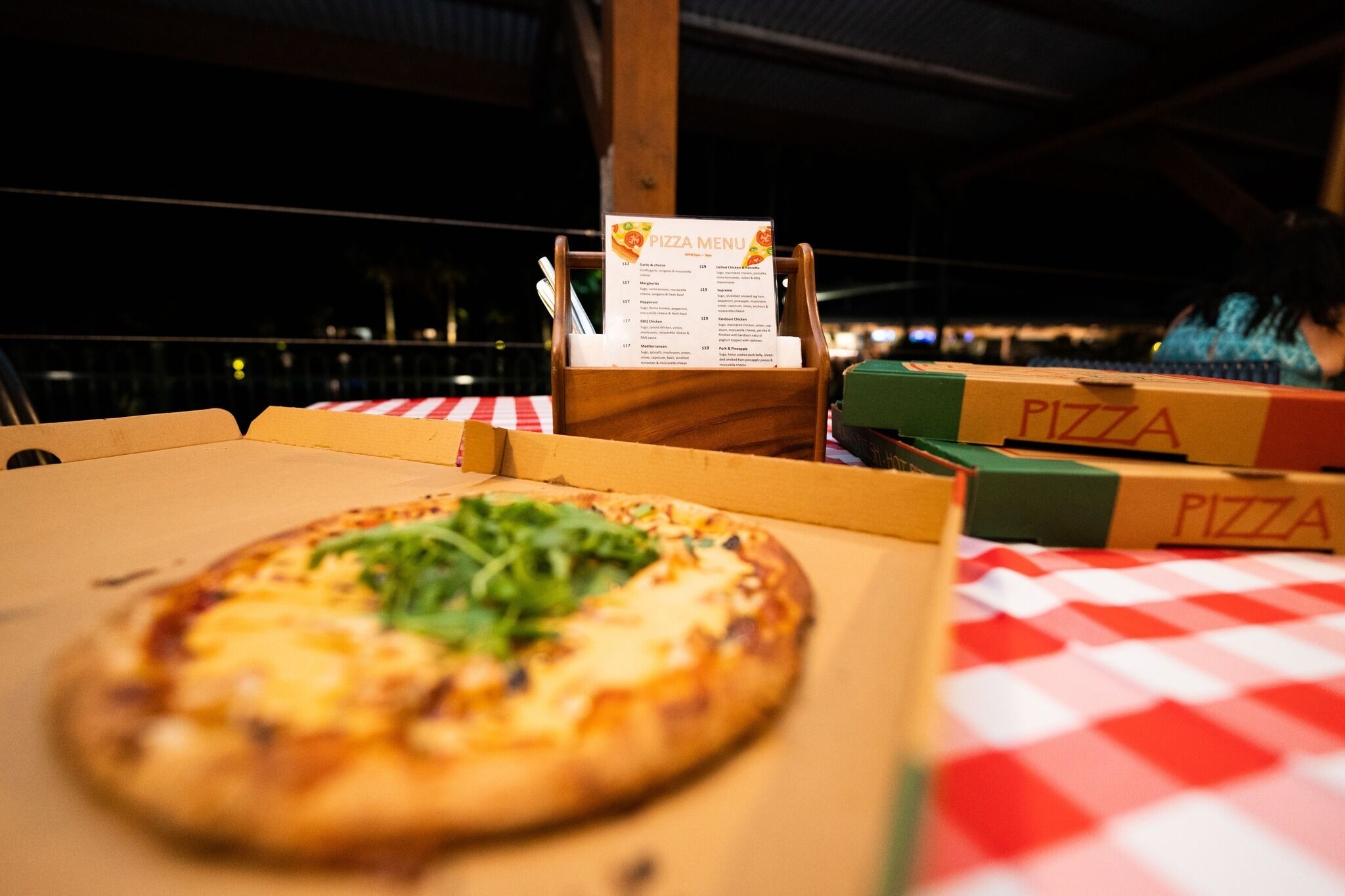 Una caja de pizza con un menú encima, sobre una mesa con mantel a cuadros rojo y blanco.