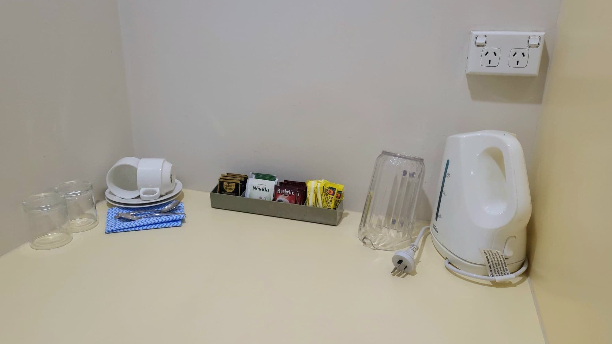 Een kitchenette met een koffiezetapparaat, een glazen pot, een doos koffiekapsules en een stopcontact.
