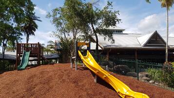 A playground with a yellow slide and a green slide, surrounded by trees and a building with a metal roof.