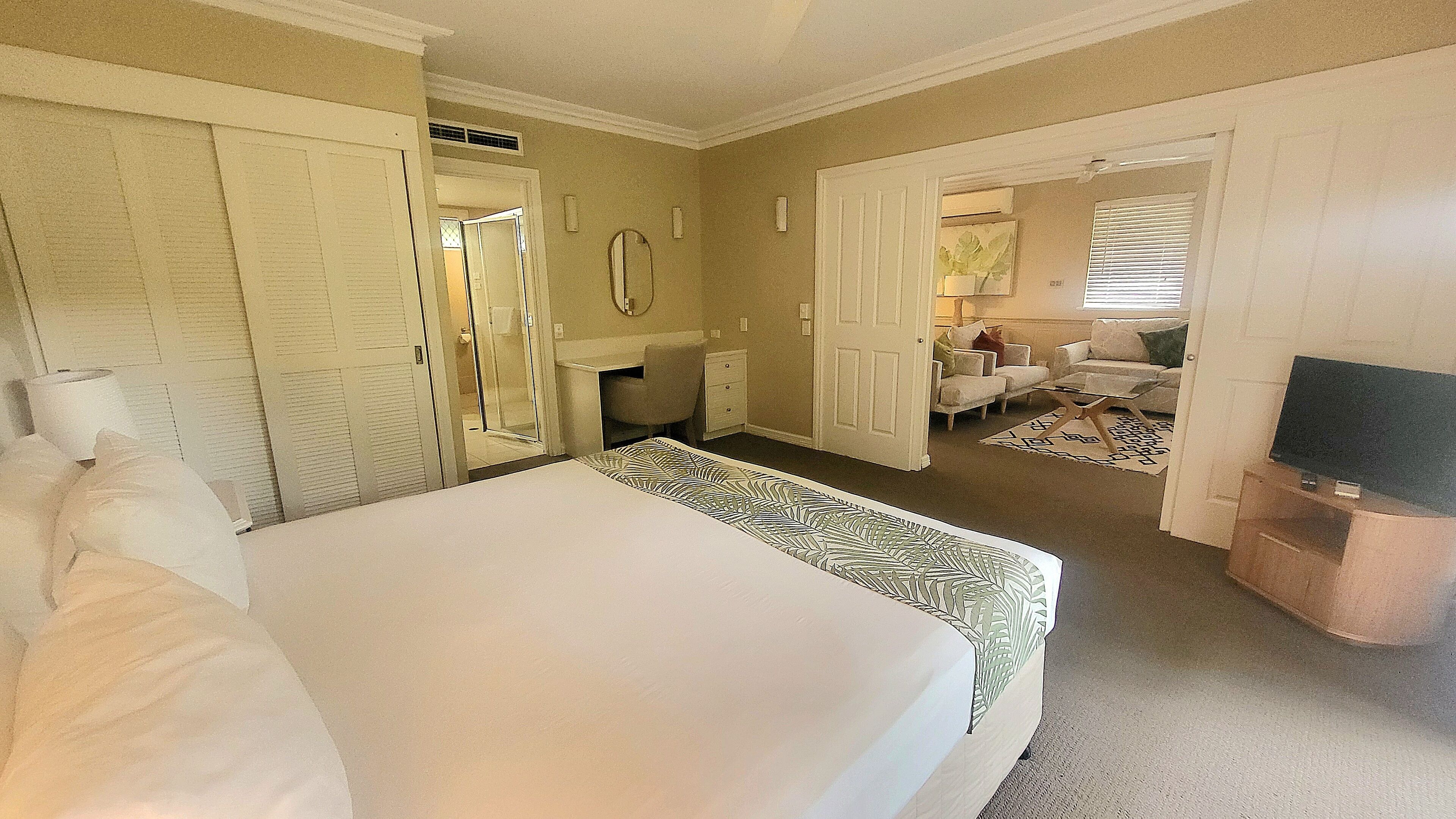 A hotel room with a large bed, a desk, a chair, a TV, and a sofa in the adjacent living area.