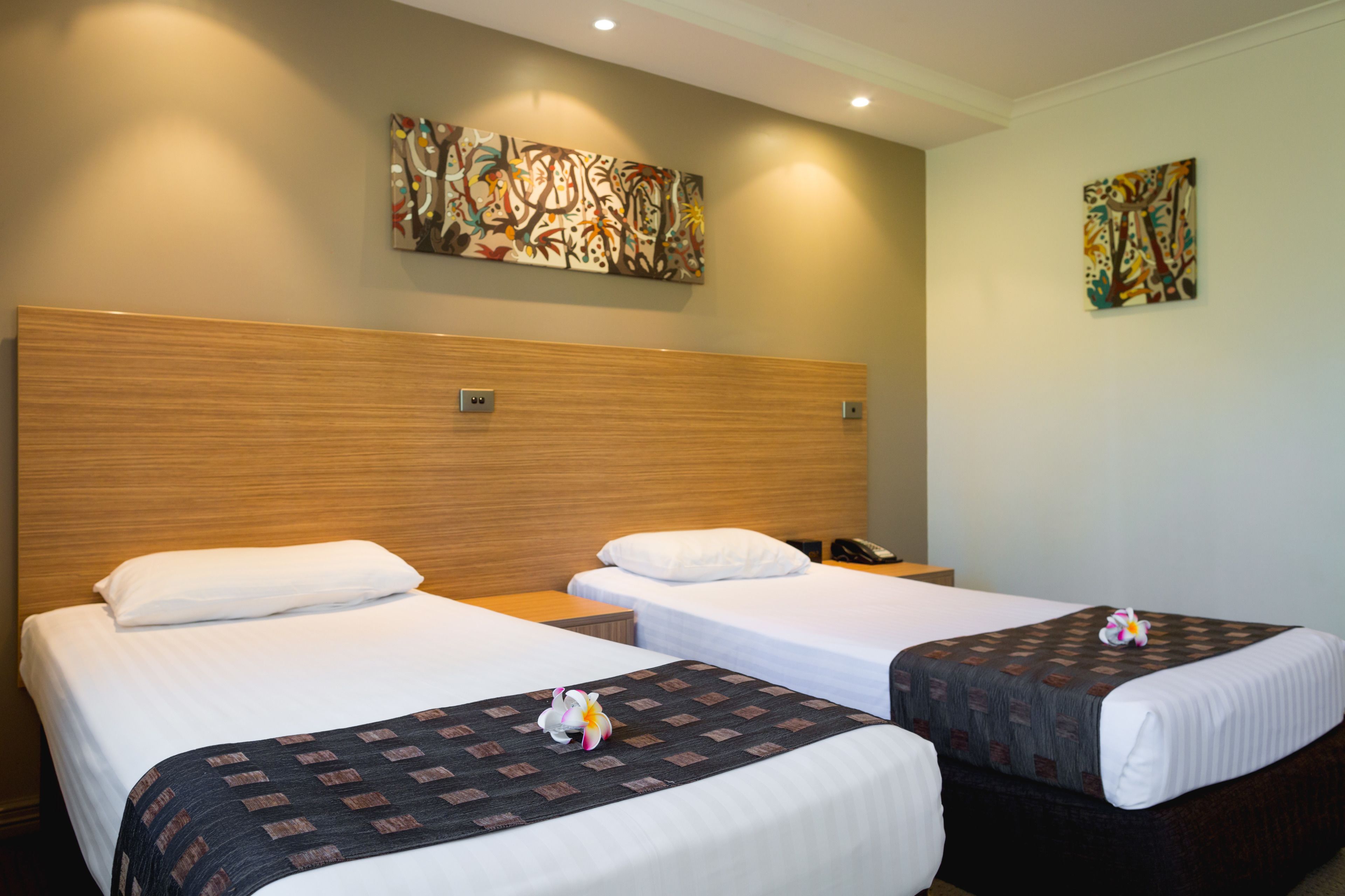 A hotel room with two beds, each with a pillow and a blanket. There are two paintings on the walls and a telephone on the nightstand.