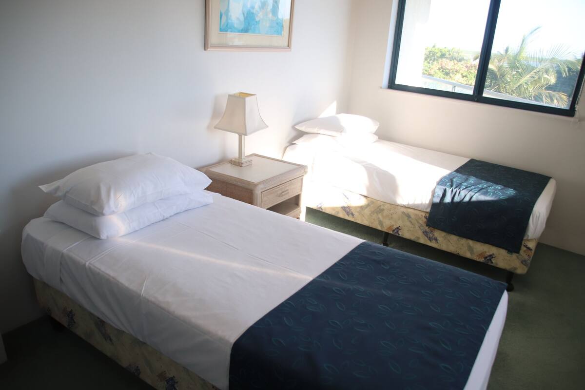 A room with two beds, one with a dark blue bedspread and the other with white bedding. There is a nightstand with a lamp and a framed picture on the wall.