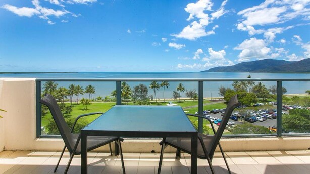 A balcony with a table and chairs, overlooking a parking area and a body of water with palm trees and mountains in the distance. - 181 The Esplanade (Cairns North)
