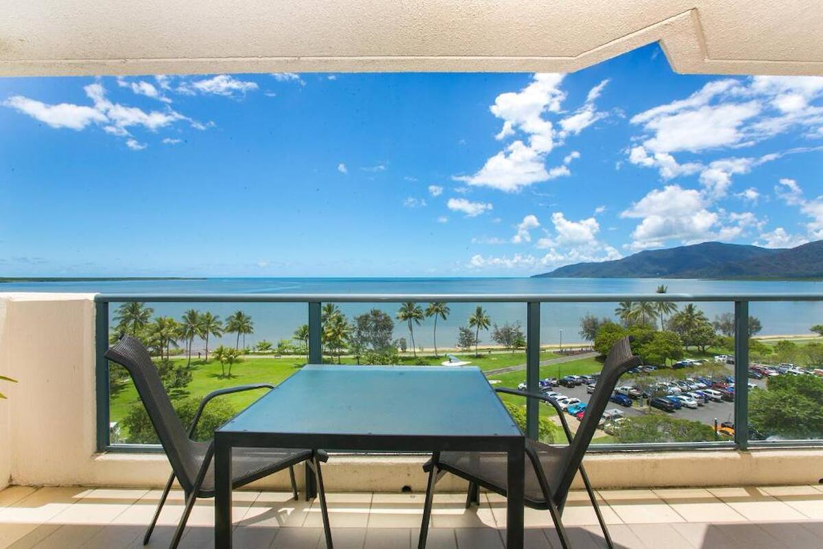 A balcony with a table and chairs, overlooking a parking area and a body of water with palm trees and mountains in the distance.