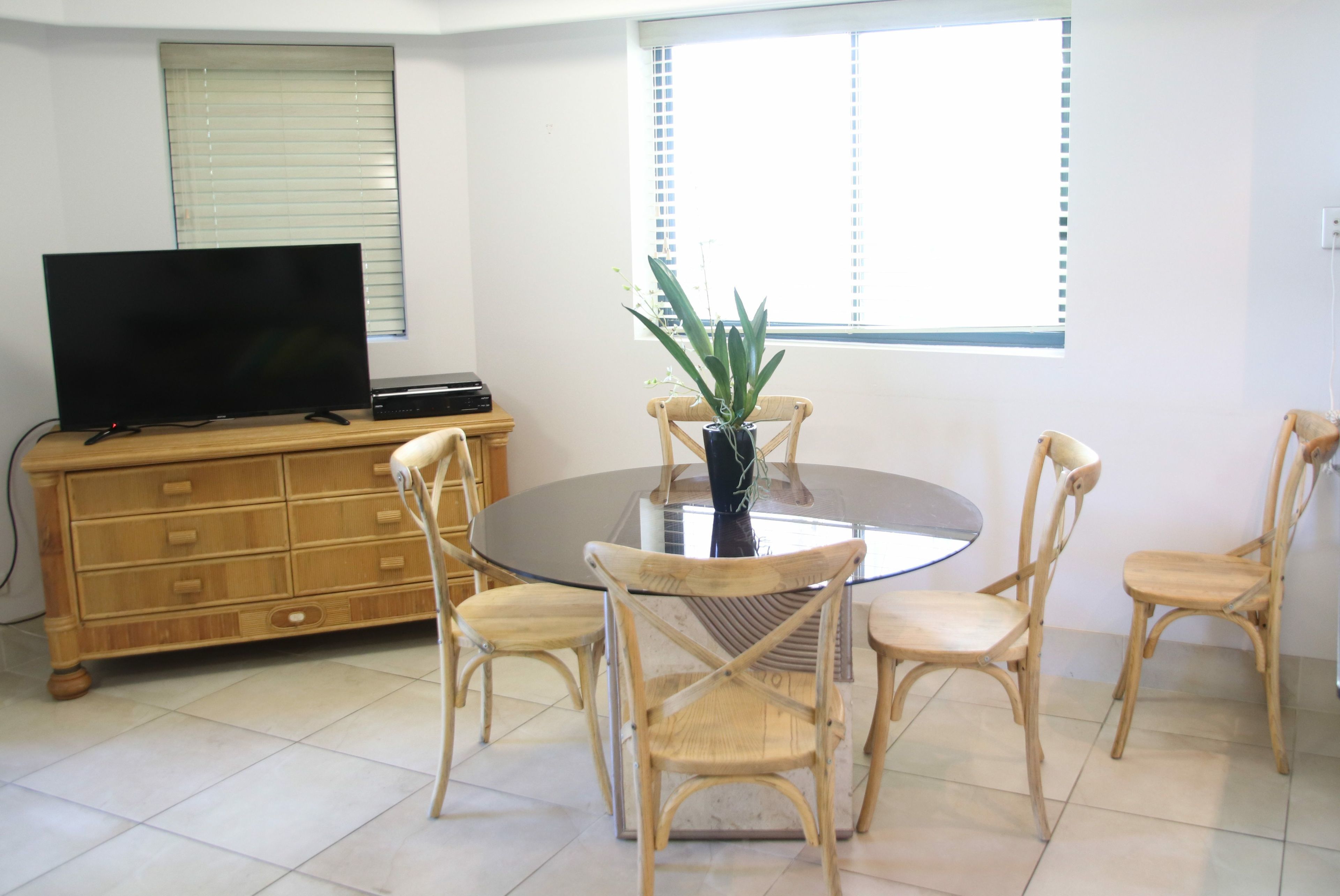A dining area with a round glass table, four wooden chairs, a television on a wooden cabinet, and a window with blinds.