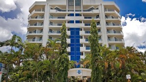 A multi-story hotel with a central blue glass facade, surrounded by palm trees. - 181 The Esplanade (Cairns North)
