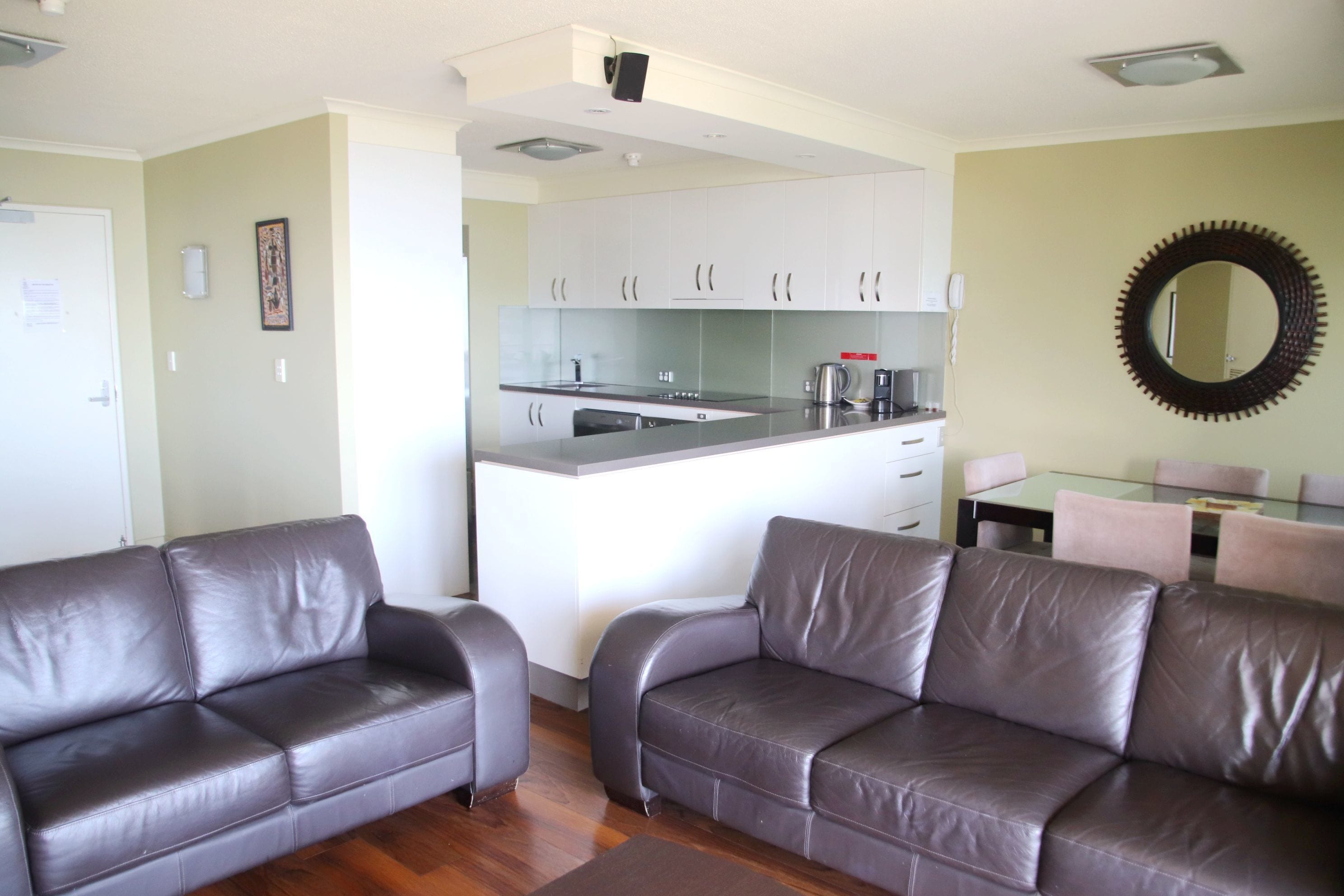 A modern living room with two leather sofas, a dining table with chairs, a kitchen area with white cabinets, and a large mirror on the wall.