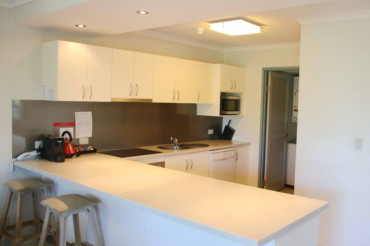 A modern kitchen with white cabinets, a built-in microwave, a kettle, and a coffee maker. There are two bar stools and a note on the counter.