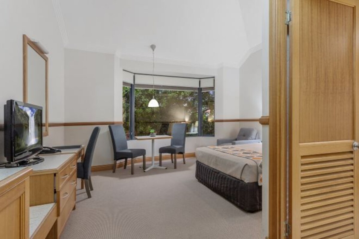 Une chambre d’hôtel équipée d’un lit, d’un bureau, de chaises, d’une télévision et d’une fenêtre donnant sur un paysage verdoyant.