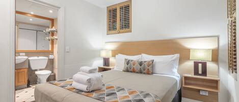 A hotel room with a large bed, two bedside tables with lamps, a headboard, a mirror, a toilet, and a shower area.