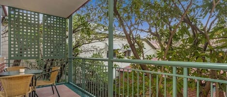 A balcony with a green metal railing, a wicker table with chairs, and a view of trees and buildings.