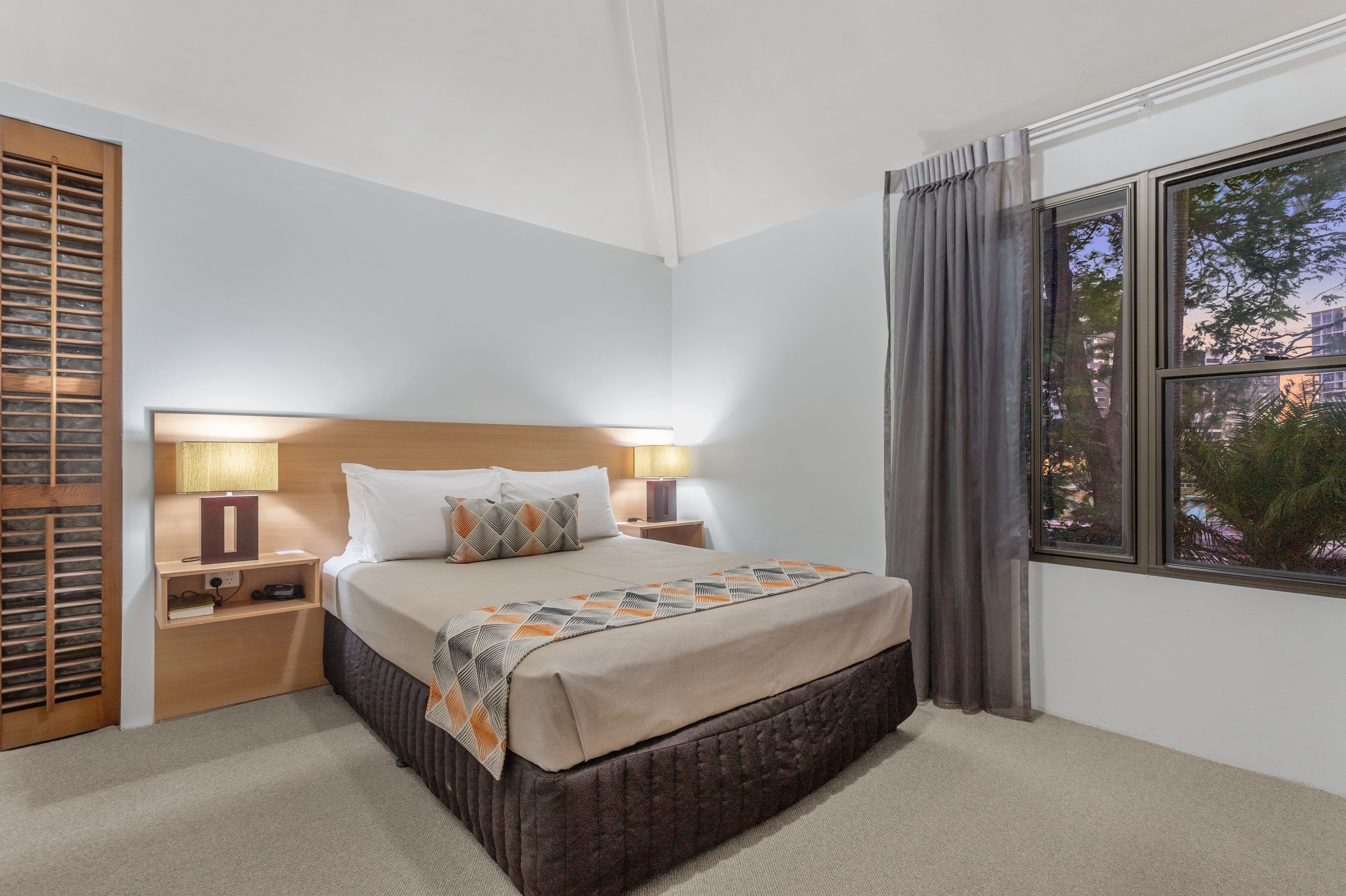 Une chambre d’hôtel avec un grand lit, deux tables de chevet, une commode, une fenêtre avec des rideaux et une vue sur les bâtiments environnants.