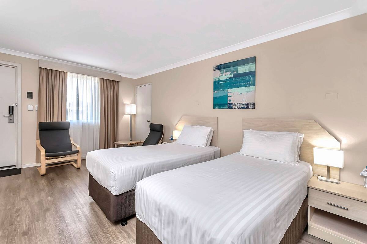 A hotel room with two single beds, a chair, a desk, a lamp, and a painting on the wall.