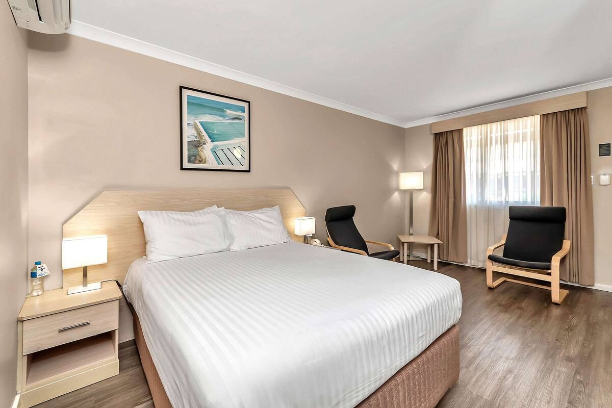 A hotel room with a large bed, two chairs, a nightstand, a lamp, and a painting of a beach scene.
