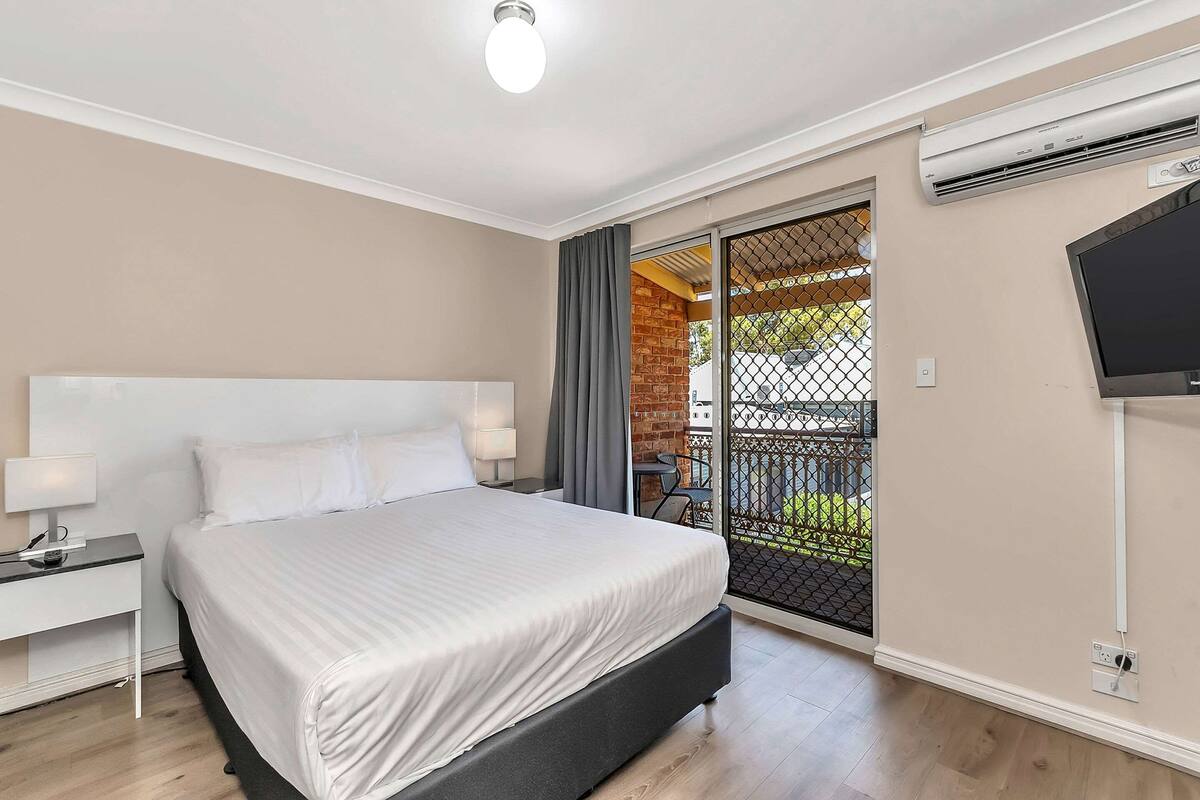A hotel room with a bed, bedside tables, a TV, and an air conditioning unit.