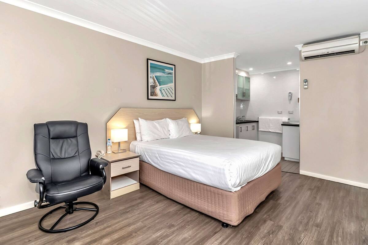 A hotel room with a bed, a desk with a chair, a small kitchenette, and an air conditioning unit.