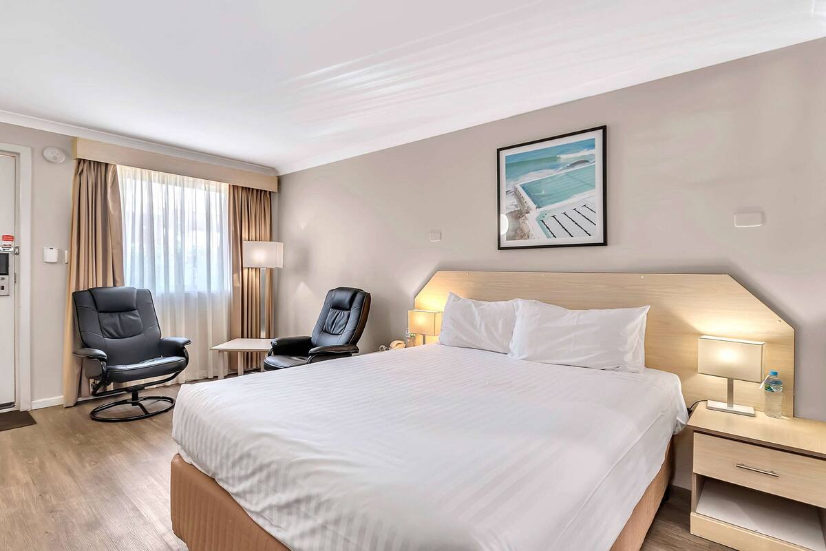 A hotel room with a large bed, two chairs, a small table, a nightstand with a lamp, and a framed picture on the wall.